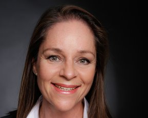 Porträt einer Frau mit langen, braunen Haaren, die ein weißes Hemd und einen schwarzen Blazer trägt. Sie lächelt freundlich in die Kamera.