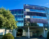 Moderne Gebäude mit großen Glasfenstern und einem begrünten Eingangsbereich. Ein gepflasterter Weg führt zur Eingangstür, umgeben von Bäumen und Sträuchern. Der Himmel ist klar und blau.