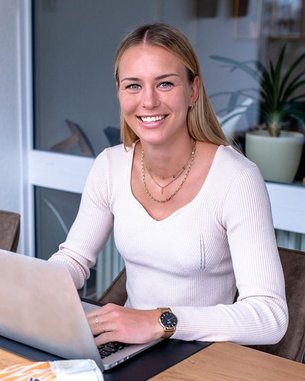 Eine Frau sitzt an einem Tisch mit einem Laptop, einem Fußball und einer Tasse. Sie trägt einen hellen Pullover und hat Handschuhe für Torhüter neben sich.