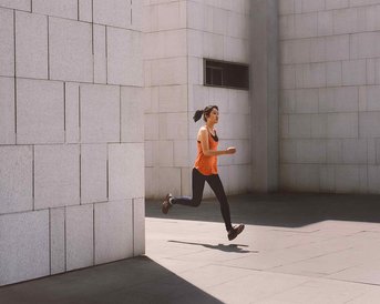 Eine Frau in einem orangefarbenen Top joggt auf einem gepflasterten Platz zwischen modernen Wänden.