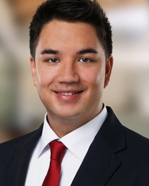 SRH Fernhochschule | Alumni-Portrait des Monats Ein junger Mann mit dunklen Haaren trägt einen Anzug und eine rote Krawatte. Er lächelt und steht vor einem unscharfen, modernen Hintergrund.