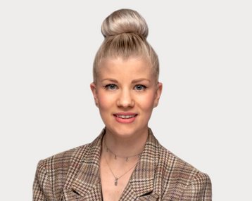 Eine Person mit blonden Haaren im Dutt trägt einen karierten Blazer und lächelt in die Kamera. Der Hintergrund ist neutral.