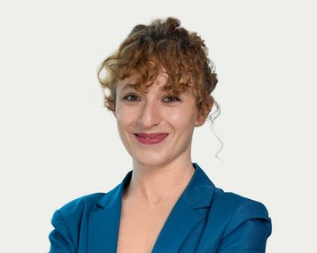 Eine lächelnde Frau mit lockigem Haar trägt einen blauen Blazer und steht mit verschränkten Armen vor einem hellen Hintergrund.