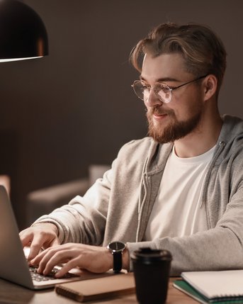 Online-Abendstudium Soziale Arbeit Ein junger Mann mit Brille sitzt an einem Tisch und arbeitet an einem Laptop. Er trägt einen grauen Hoodie und hat eine Tasse Kaffee sowie Notizmaterial neben sich. Die Beleuchtung ist gedämpft.