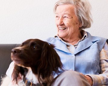 Eine ältere Frau sitzt auf einem Sofa und lächelt, während ein Hund neben ihr liegt. Sie trägt eine hellblaue Weste und hat graue Haare.