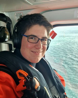 Eine Person mit Brille und Kopfhörern sitzt in einem Hubschrauber, trägt eine orangefarbene Sicherheitsweste und lächelt. Im Hintergrund ist Wasser und eine Windkraftanlage sichtbar.