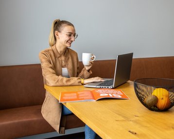 Eine Frau sitzt an einem Tisch, arbeitet am Laptop und hält eine Tasse in der Hand. Auf dem Tisch liegt eine Zeitschrift und eine Schüssel mit Obst.