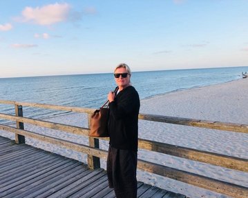 Eine Person mit Sonnenbrille steht auf einem Holzsteg am Strand, hält eine Tasche und blickt zur Kamera. Im Hintergrund ist der Ozean und ein sandiger Strand zu sehen.