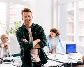 Ein Mann mit lässiger Kleidung steht lächelnd im Vordergrund eines modernen Büros, während zwei Personen im Hintergrund an einem Tisch arbeiten. Eine Frau beugt sich über einen Computer, während ein Mann am Tisch sitzt.