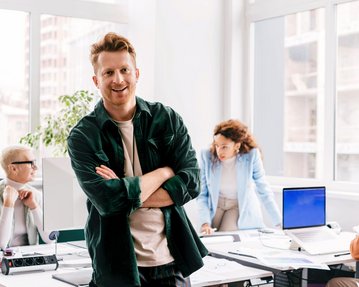 Kommunikation in Organisationskrisen Ein Mann mit lässiger Kleidung steht lächelnd im Vordergrund eines modernen Büros, während zwei Personen im Hintergrund an einem Tisch arbeiten. Eine Frau beugt sich über einen Computer, während ein Mann am Tisch sitzt.