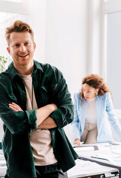Ein Mann mit lässiger Kleidung steht lächelnd im Vordergrund eines modernen Büros, während zwei Personen im Hintergrund an einem Tisch arbeiten. Eine Frau beugt sich über einen Computer, während ein Mann am Tisch sitzt.