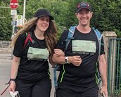 Ein Mann und eine Frau in sportlicher Kleidung mit Rucksäcken gehen zusammen auf einem Gehweg. Beide tragen T-Shirts mit dem Aufdruck "Neckarsteig 2022".