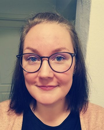 Eine Frau mit kurzen, dunklen Haaren und Brille lächelt in die Kamera. Sie trägt ein schwarzes T-Shirt und eine rosafarbene Strickjacke. Der Hintergrund zeigt eine Wand und eine Tür.