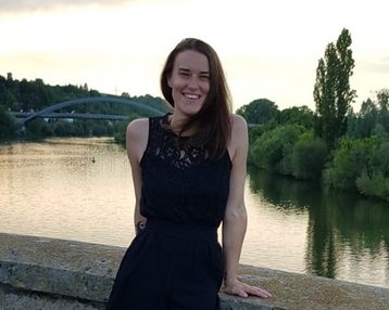 Eine Frau in schwarzem Outfit steht lächelnd an einer Steinmauer am Fluss, mit Bäumen und einer Brücke im Hintergrund. Der Himmel ist bewölkt und das Wasser spiegelt die Umgebung wider.