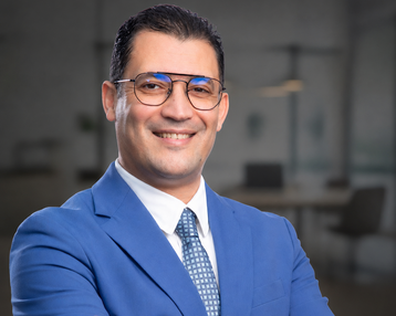 Ein Mann in einem blauen Anzug mit Brille lächelt, während er mit verschränkten Armen vor einem modernen Bürohintergrund steht.