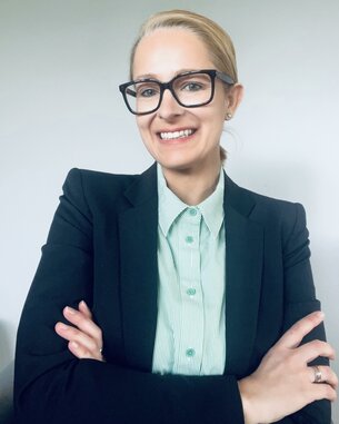 Eine Frau mit blonden Haaren trägt eine schwarze Blazerjacke und ein gestreiftes Hemd. Sie lächelt und hat die Arme verschränkt.