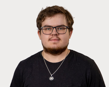 Ein junger Mann mit Brille und Bart trägt ein schwarzes T-Shirt und eine Halskette. Er schaut direkt in die Kamera.