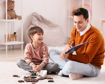 Ein Erwachsener und ein Kind sitzen auf dem Boden und spielen mit Buchstaben aus Holz. Der Erwachsene schreibt in ein Notizbuch.