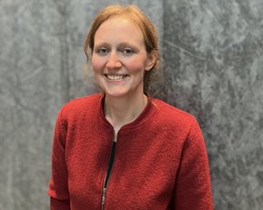 Eine Person mit blonden Haaren trägt eine rote Jacke und lächelt vor einer grauen Wand.