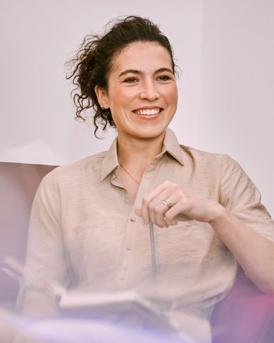 Eine Frau mit lockigem Haar sitzt entspannt in einem Sessel, hält einen Stift in der Hand und lächelt. Sie trägt ein beiges Hemd und hat ein Notizbuch auf ihrem Schoß.