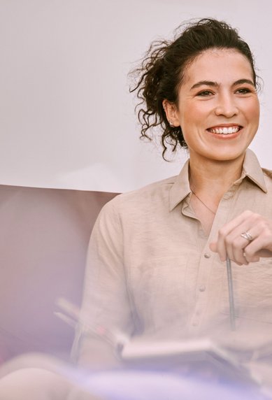 Eine Frau mit lockigem Haar sitzt entspannt in einem Sessel, hält einen Stift in der Hand und lächelt. Sie trägt ein beiges Hemd und hat ein Notizbuch auf ihrem Schoß.