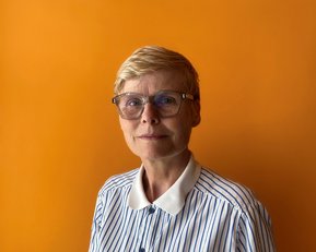 Eine Frau mit kurzen, blonden Haaren und einer gestreiften Bluse steht vor einem orangefarbenen Hintergrund. Sie trägt eine modische Brille.