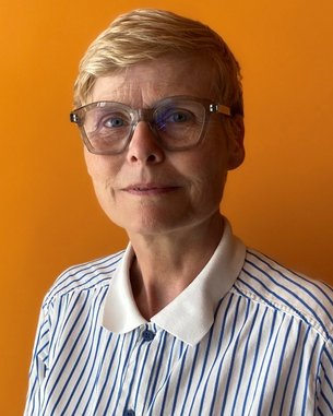 Eine Frau mit kurzen, blonden Haaren und einer gestreiften Bluse steht vor einem orangefarbenen Hintergrund. Sie trägt eine modische Brille.