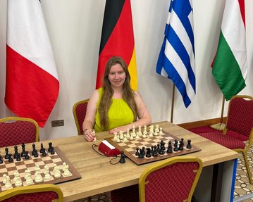 Eine Schachspielerin sitzt an einem Tisch mit zwei Schachbrettern, umgeben von Flaggen verschiedener Länder.