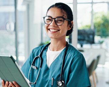 Eine lächelnde Krankenschwester in grüner Arbeitskleidung hält ein Tablet in der Hand. Im Hintergrund sind weitere medizinische Fachkräfte zu sehen.