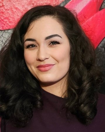 Eine Frau mit lockigem, dunklem Haar und einem lila Oberteil lächelt in die Kamera, vor einem bunten Hintergrund.