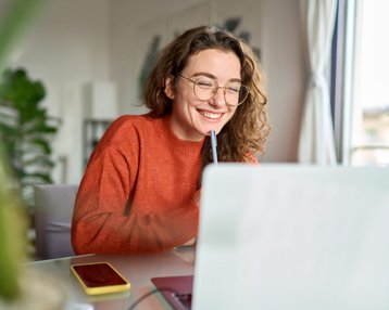 Eine lächelnde Frau mit Brille sitzt an einem Tisch, arbeitet am Laptop und hält einen Stift in der Hand. Ein Smartphone liegt daneben.