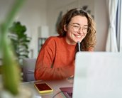 Eine lächelnde Frau mit Brille sitzt an einem Tisch, arbeitet am Laptop und hält einen Stift in der Hand. Ein Smartphone liegt daneben.