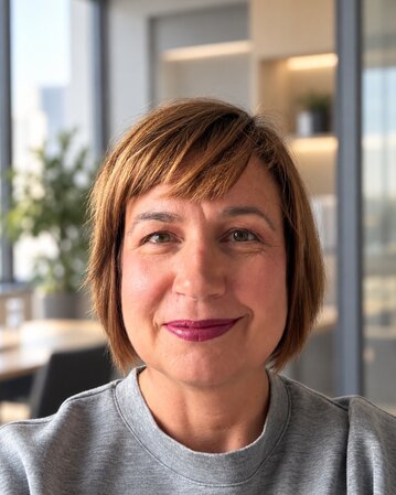 Frau mit schulterlangen, braunen Haaren und lila Lippenstift, lächelt in einem modernen Büro mit Pflanzen im Hintergrund.