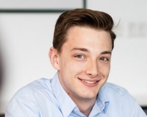 Ein junger Mann mit braunen Haaren trägt ein hellblaues Hemd und lächelt freundlich, während er an einem Tisch sitzt.