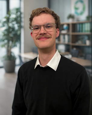 Junger Mann mit Brille und Schnurrbart, trägt einen schwarzen Pullover und ein weißes Hemd, steht in einem modernen Büro mit Pflanzen im Hintergrund.