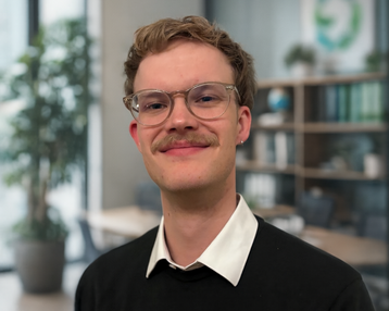 Junger Mann mit Brille und Schnurrbart, trägt einen schwarzen Pullover und ein weißes Hemd, steht in einem modernen Büro mit Pflanzen im Hintergrund.