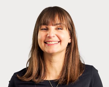 Eine lächelnde Frau mit langen, braunen Haaren trägt ein schwarzes Oberteil. Der Hintergrund ist neutral und hell.