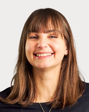 Eine lächelnde Frau mit langen, braunen Haaren trägt ein schwarzes Oberteil. Der Hintergrund ist neutral und hell.