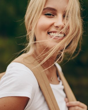 Junge Frau mit blonden Haaren und lächelndem Gesicht, trägt ein weißes T-Shirt und eine beige Tasche. Der Hintergrund zeigt verschwommenes Grün.