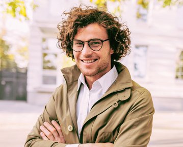Ein junger Mann mit lockigem Haar und Brille lächelt, während er in einem beigen Mantel mit verschränkten Armen steht. Im Hintergrund sind unscharfe Gebäude und Bäume sichtbar.