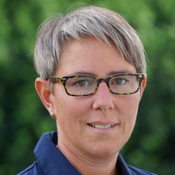 Eine Person mit kurzen, grauen Haaren und einer blauen Bluse trägt eine Brille mit einem tortoiseshell Muster. Der Hintergrund zeigt unscharfe grüne Pflanzen.