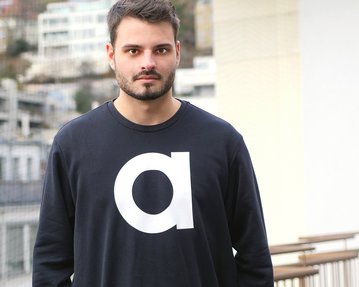 Ein Mann mit Bart trägt ein schwarzes Sweatshirt mit einem großen weißen Buchstaben "a" auf der Vorderseite. Er steht auf einem Balkon mit einer urbanen Kulisse im Hintergrund.