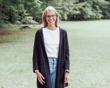 Eine Frau steht auf einer Wiese, trägt eine schwarze Strickjacke, ein weißes T-Shirt, enge Jeans und schwarze Stiefel. Sie hat kurze, blonde Haare und eine Brille. Im Hintergrund sind Bäume zu sehen.