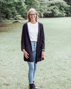 Eine Frau steht auf einer Wiese, trägt eine schwarze Strickjacke, ein weißes T-Shirt, enge Jeans und schwarze Stiefel. Sie hat kurze, blonde Haare und eine Brille. Im Hintergrund sind Bäume zu sehen.