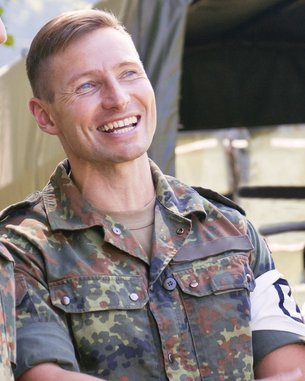 Drei Soldaten in Uniform stehen im Freien und unterhalten sich. Sie lächeln und wirken entspannt, während ein Zelt im Hintergrund sichtbar ist.