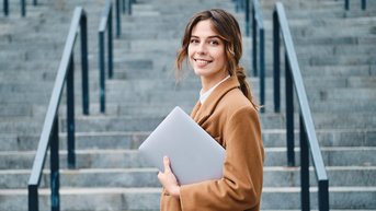 Junge Frau mit braunem Mantel steht an einer Treppe und hält einen Laptop in der Hand. Sie lächelt in die Kamera.