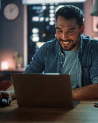 Ein Mann sitzt an einem Tisch und lächelt, während er auf einen Laptop schaut. Neben ihm stehen eine Tasse und eine Kerze. Im Hintergrund sind Bücherregale und Fenster mit Stadtansicht zu sehen.