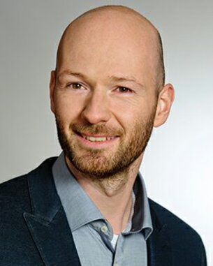 Ein Mann mit Glatze und Bart lächelt in einem Anzug mit Hemd. Der Hintergrund ist neutral und hell.