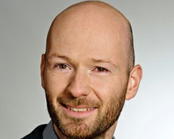Ein Mann mit Glatze und Bart lächelt in einem Anzug mit Hemd. Der Hintergrund ist neutral und hell.