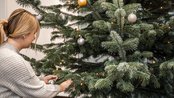 Eine Person kniet neben einem Weihnachtsbaum, während sie Lichterketten anbringt. Der Baum ist mit bunten Kugeln geschmückt und steht auf einem grünen Baumständer.